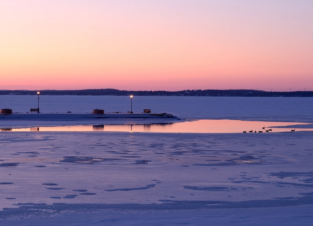 Winter evening landscape with after sunset sky reflects in the open water spot on a frozen Mendota lake with silhouettes of geese. Glowing lanterns along a pier. Tenney Park, Madison, Wisconsin