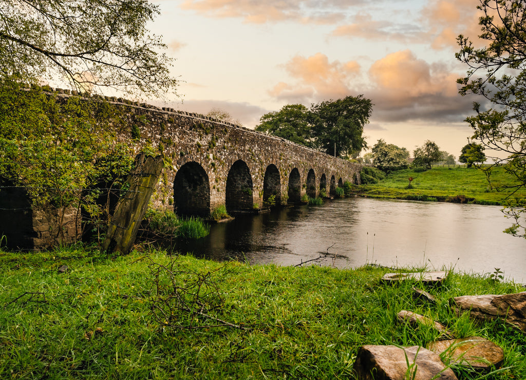 Old 12th century stone arch bridge over a river, rocks on first plane. Green fields and trees. Dramatic sky sunset. Count Meath, Ireland