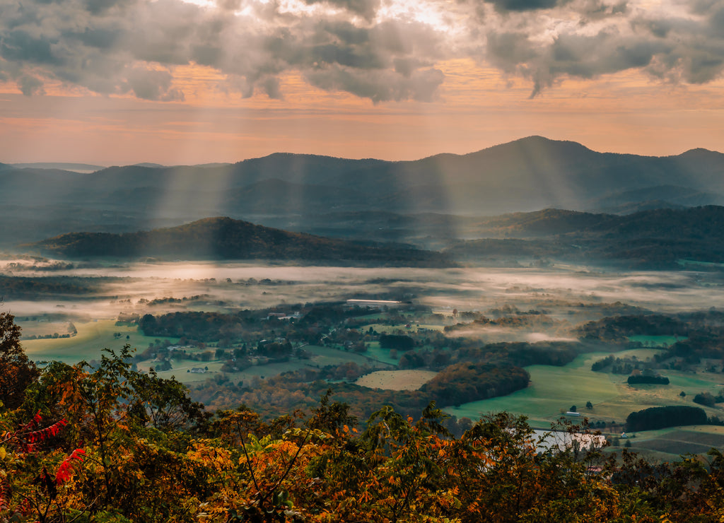 Light rays shine down on Rockfish Valley illuminating the fog and low lying clouds during a beautiful orange sunrise surrounded by colorful fall foliage, along the Blue Ridge Parkway, Virginia, USA