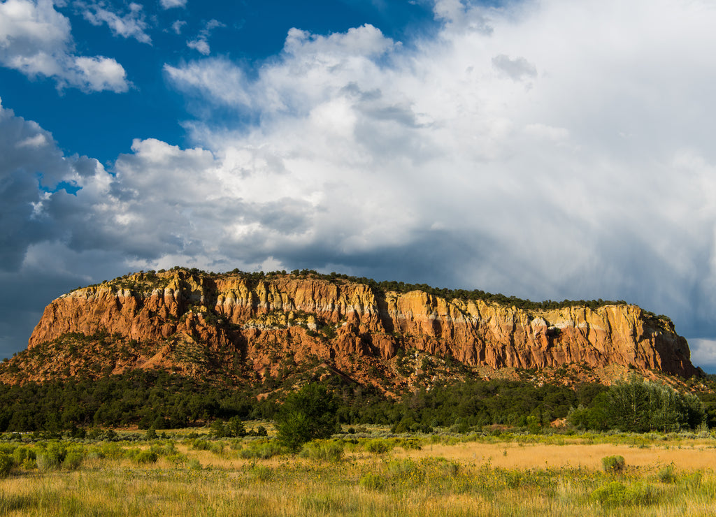 Mesa with red rock sandstone cliffs above a grassy meadow under a dramatic stormy sky in northern New Mexico