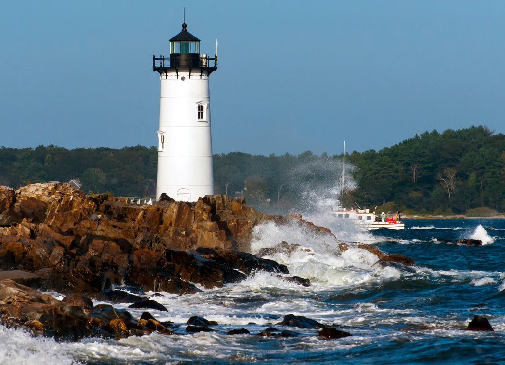Portsmouth Harbor lighthouse guides fishing boat as waves break over rocky shore during high tide in New Hampshire. The lighthouse is also referred to as Fort Constitution light