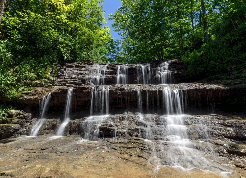 Rivulets of falling water over rock ledges are topped by emerald green leaves and a deep blue sky on a summer day at Fallsville Falls, a beautiful tiered waterfall near Hillsboro, Ohio