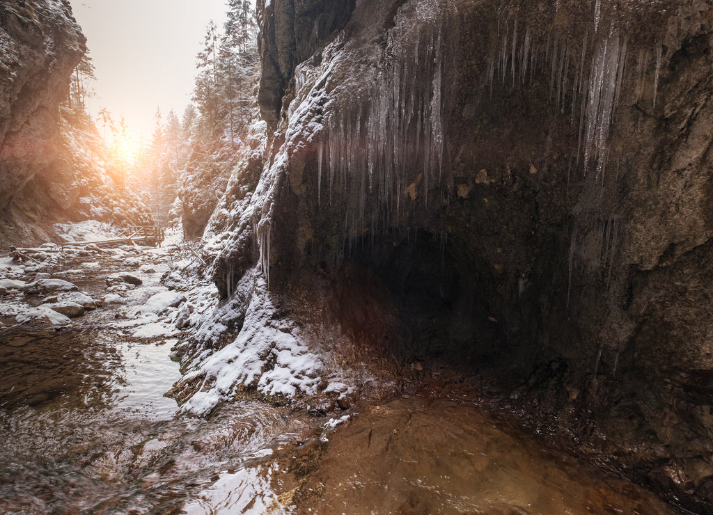 Flowing small creek running out icicle cave and washing big boulders. First sun rays lite young snow islands. Cold early winter mountain forest landscape in Slovakia Tatry mountain