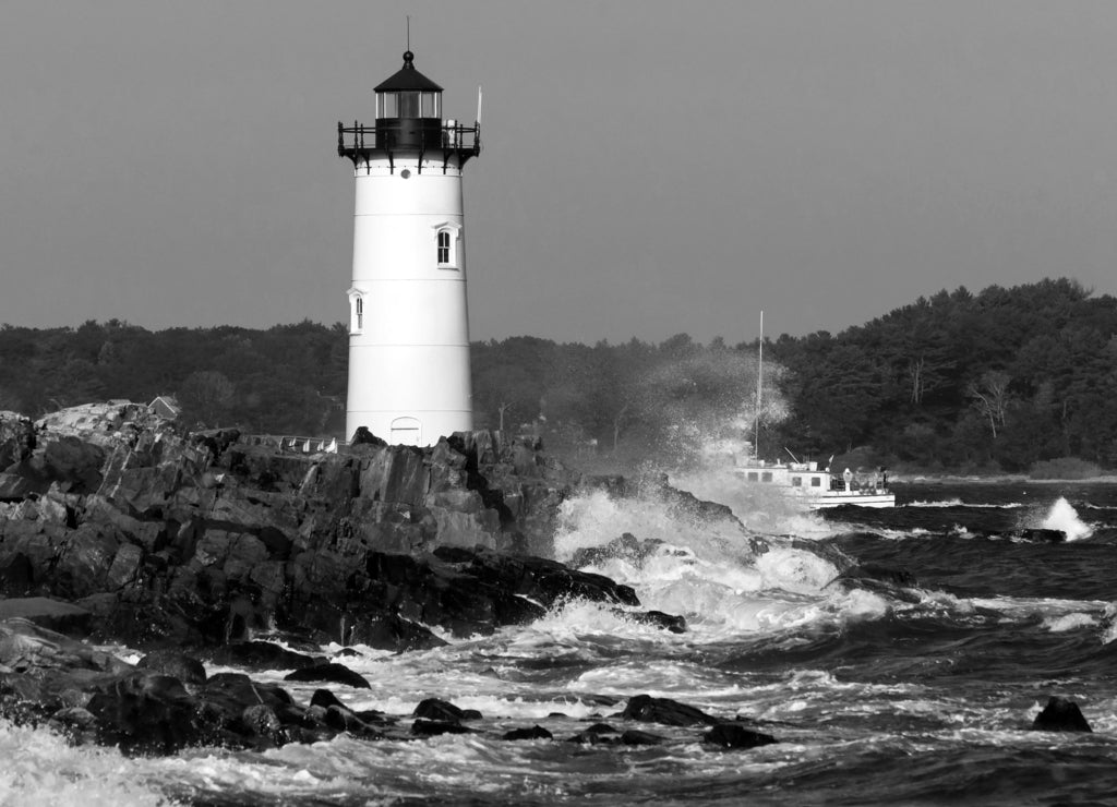Portsmouth Harbor lighthouse guides fishing boat as waves break over rocky shore during high tide in New Hampshire. The lighthouse is also referred to as Fort Constitution light in black white