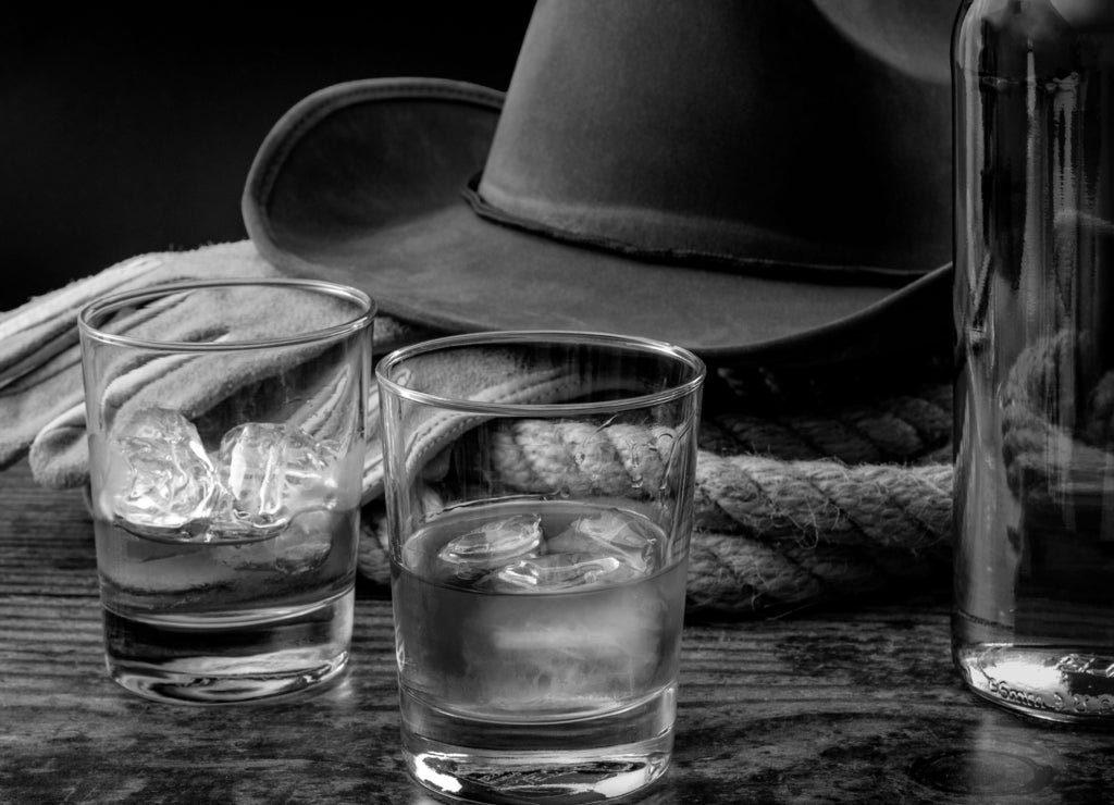 The Wild West concept theme with cowboy hat, rope lasso, leather gloves, two glasses of whiskey on the rocks and bottle of bourbon on wooden table in a vintage saloon against black background, Texas in black white