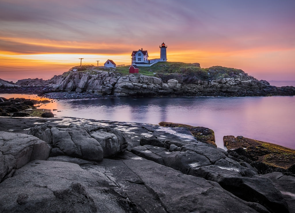 Colorful sunrise at Nubble Lighthouse, with dark rocks in the foreground and the rocky island hosting the lighthouse in the background