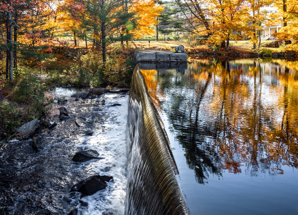 Waterfall on a lake with open floodgates framed by autumn maples lit by the sun in Vermont
