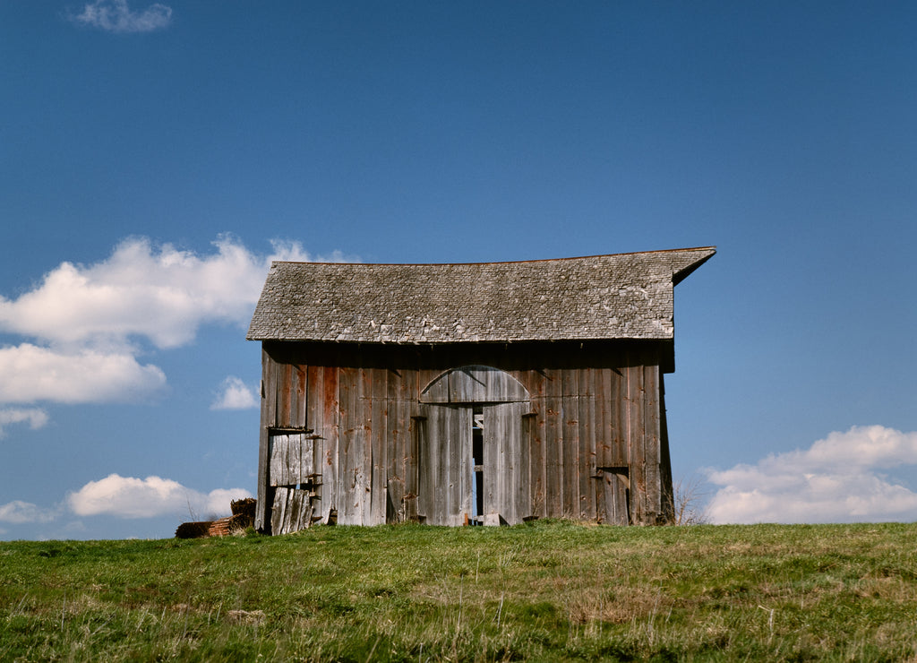 An old barn that was, at one time, red on a hillside in Adair County in Northeast Missouri with puffy clouds dotting the blue sky background. 1985