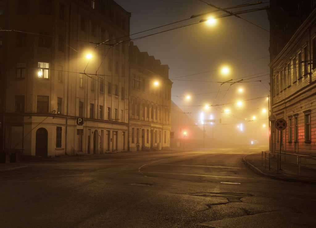 An empty illuminated asphalt road through the old historical buildings and houses in a fog at night. Street lights (lanterns) close-up. Riga, Latvia