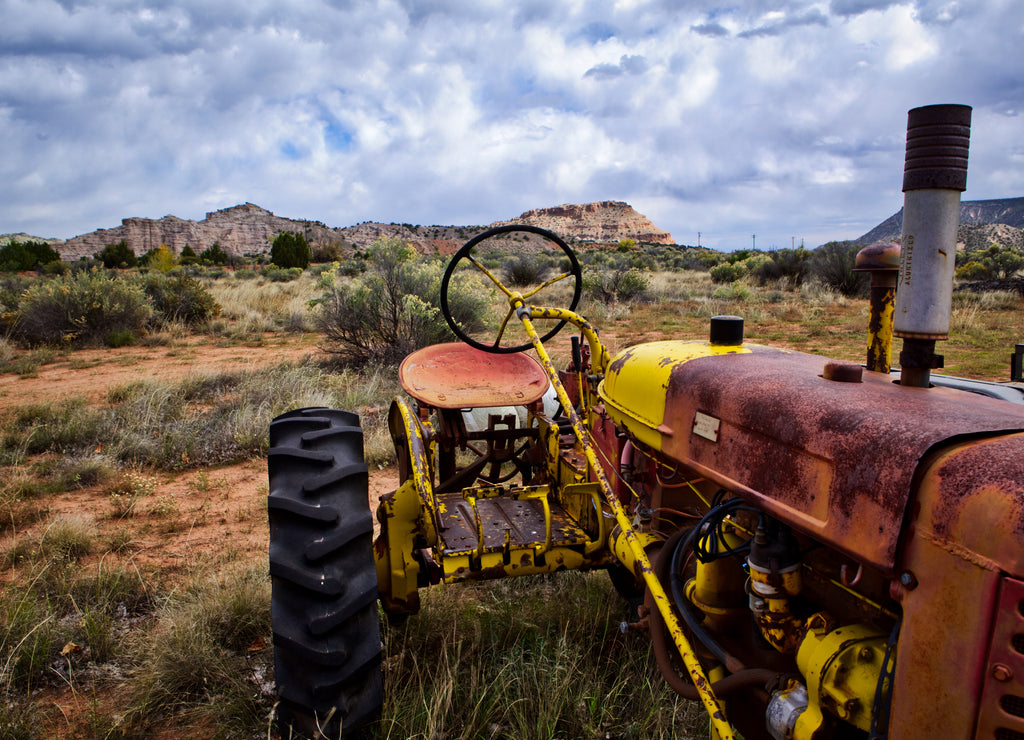 A textured and colorful old tractor sits in the New Mexico desert, with mesas and cloud-filled sky in the background