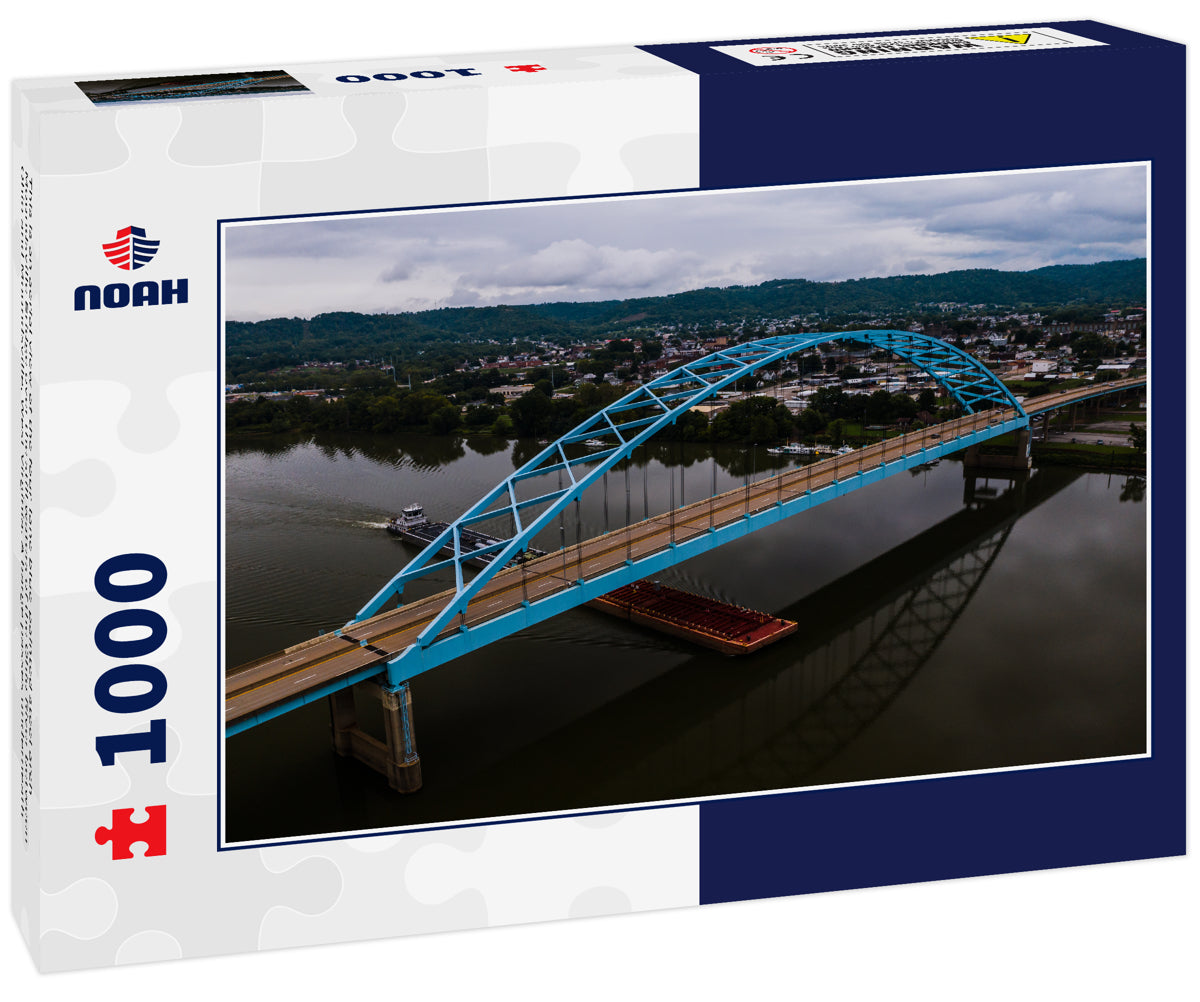 This is an aerial view of the four-lane blue-painted steel arch Moundsville Bridge over the still waters of the Ohio River between Ohio and Moundsville, West Virginia. A barge passes underneath