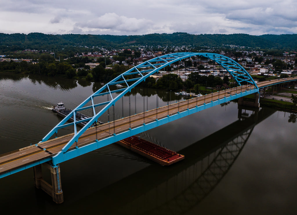 This is an aerial view of the four-lane blue-painted steel arch Moundsville Bridge over the still waters of the Ohio River between Ohio and Moundsville, West Virginia. A barge passes underneath