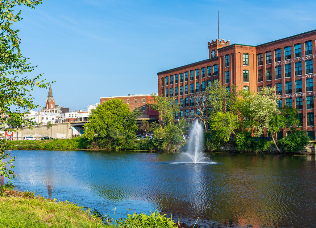 Fountain on the Nashua River against the background of a historic cotton factory building with a clock tower in the old industrial park of Nashua. New Hampshire, USA