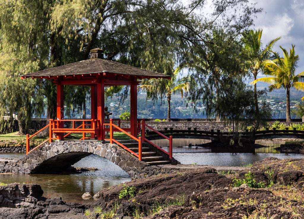 Hilo, Hawaii, USA: Closeup of Japanese black and red small bow bridge over pond in Liliuokalani Gardens. Green trees and black rocks under blue sky with big white clouds