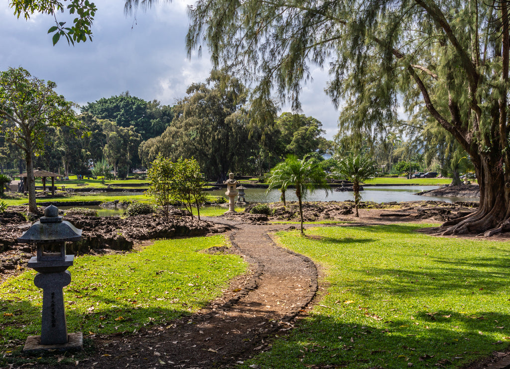 Hilo, Hawaii, USA: Path winding through green lawn among trees with Japanese lanterns on side in Liliuokalani Gardens. Sunshine but cloudscape