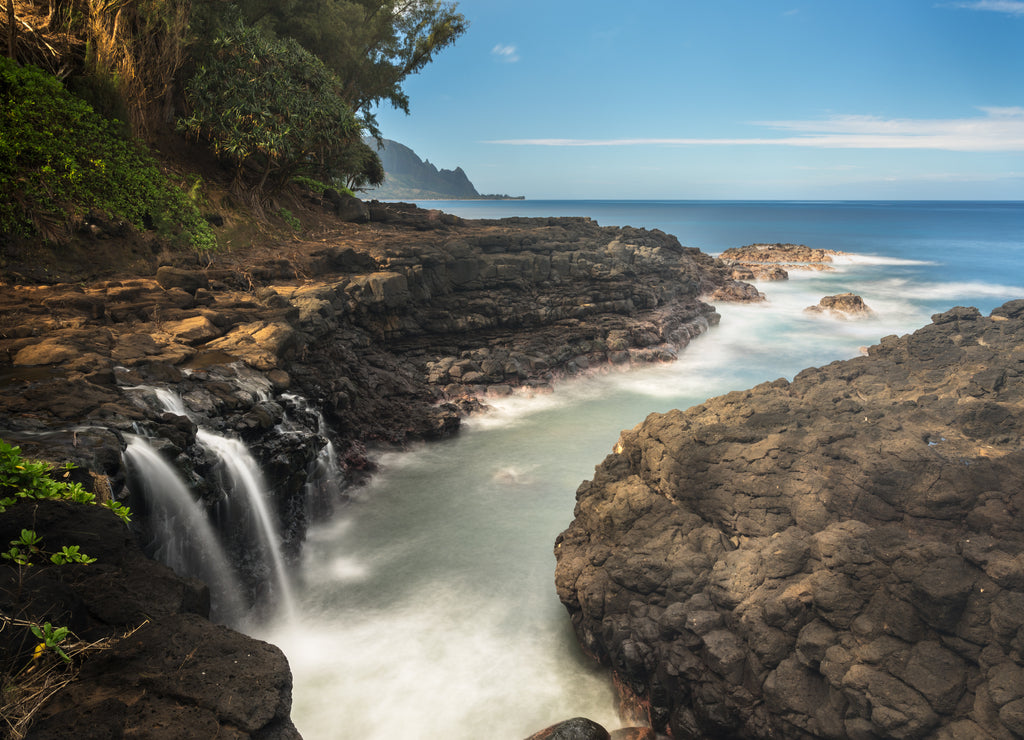 Rugged rocks around small waterfall falling into the ocean with the Na Pali coastline by Hanalei in the distance Hawaii
