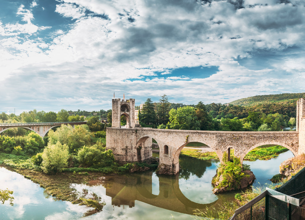 Besalu, Girona, Catalonia, Spain. Famous Landmark Old Medieval Romanesque Besalu Bridge Over The Fluvia River In Cloudy Summer Day