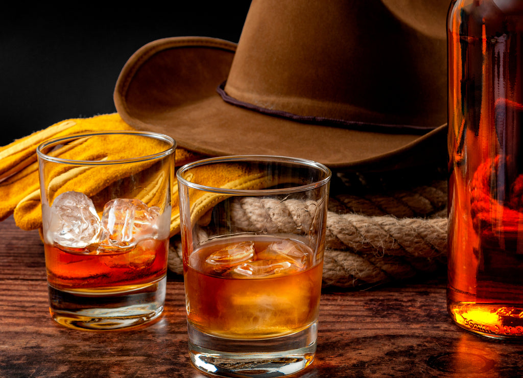 The Wild West concept theme with cowboy hat, rope lasso, leather gloves, two glasses of whiskey on the rocks and bottle of bourbon on wooden table in a vintage saloon against black background, Texas