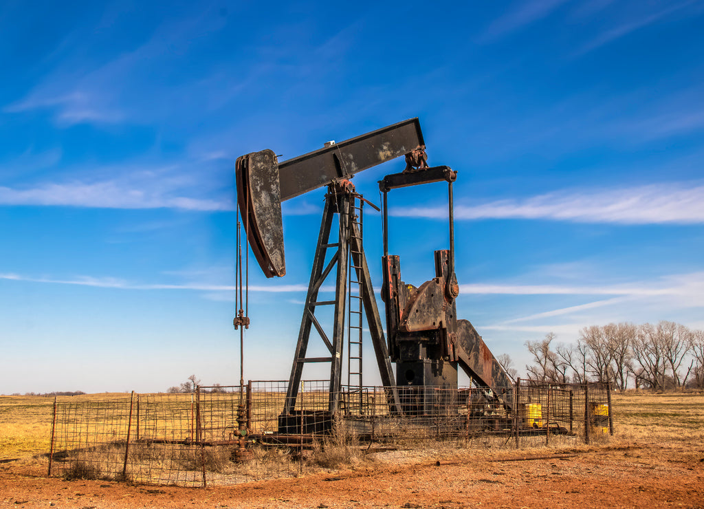 Large old rusty oil well pump jack surrounded by cattle panel fence out in field, Oklahoma