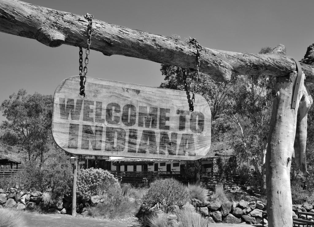 Old wood signboard with text " welcome to indiana" hanging on a branch in black white