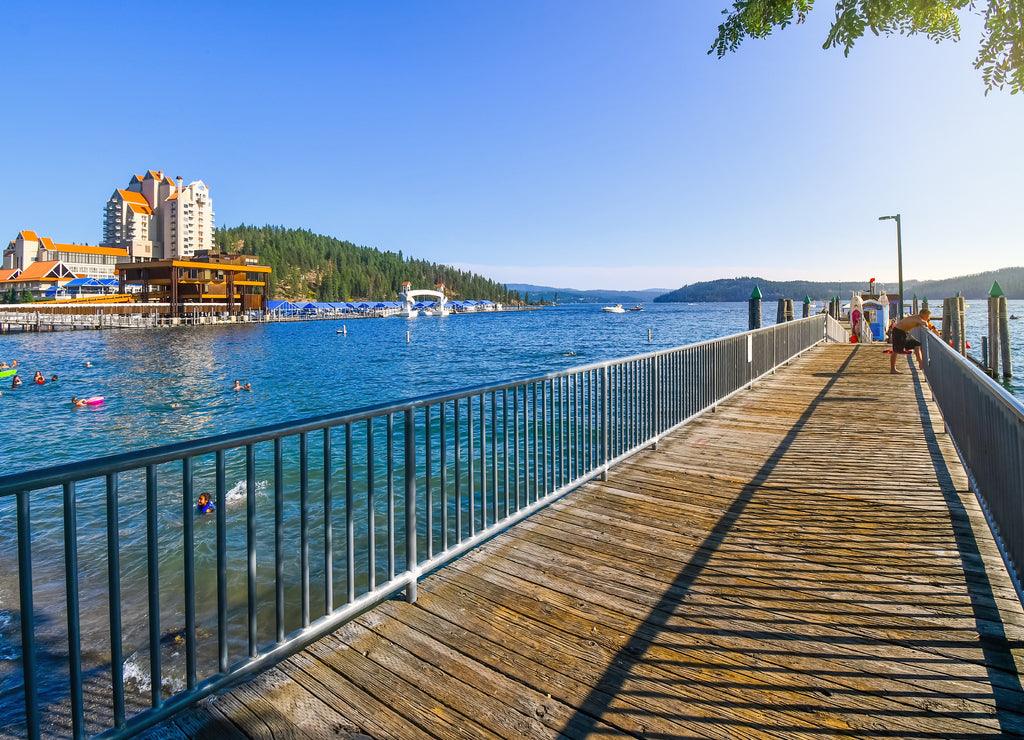 A young boy leans over the dock edge at afternoon at a lakefront wooden dock with a hotel resort and mountain behind in Coeur d'Alene Idaho USA