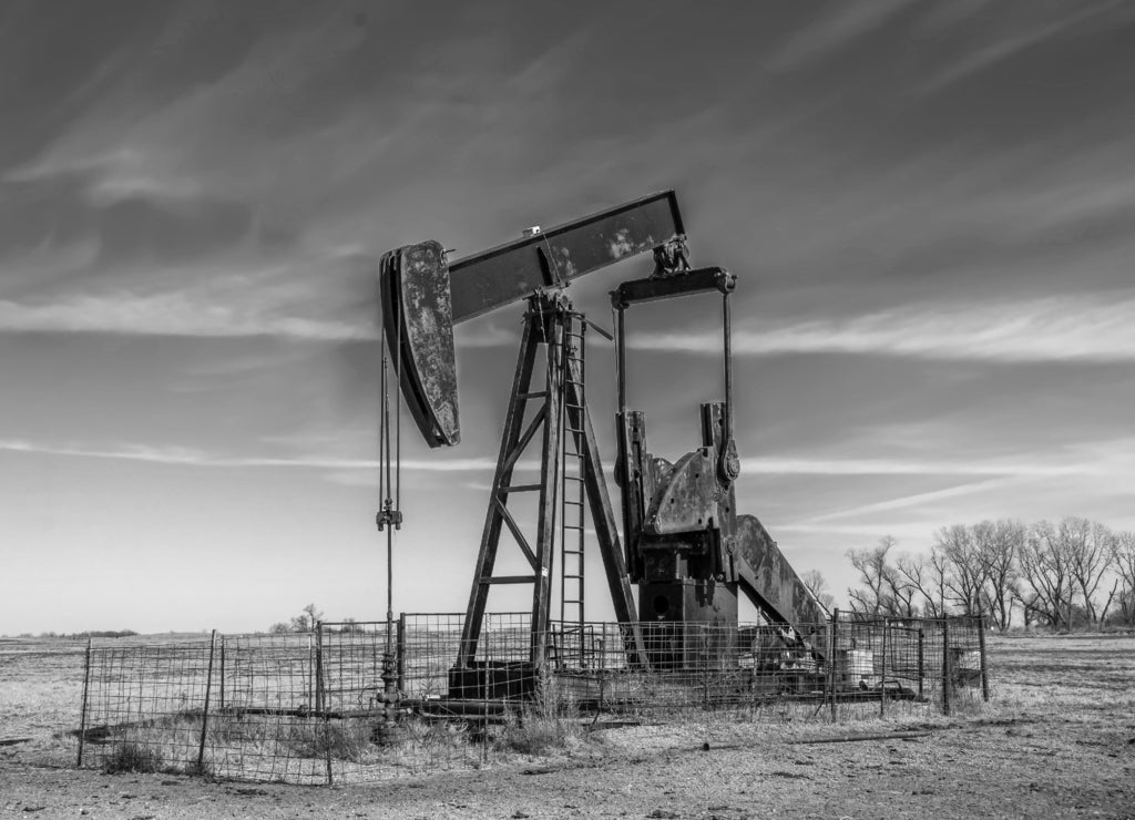 Large old rusty oil well pump jack surrounded by cattle panel fence out in field, Oklahoma in black white