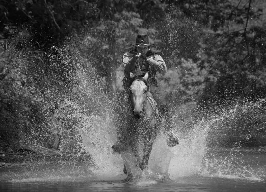 A vintage action shot of a cowboy riding a horse, wading through the water and holding a rifle, Texas in black white