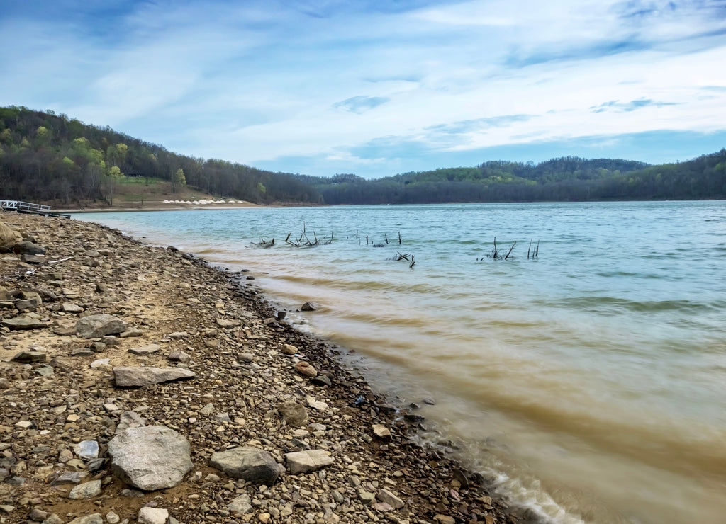 Tygart Lake in Tygart Lake State Park in West Virginia, in the spring with the shoreline filled with sand and rocks leading to the wavy water with the trees and sky filled with clouds
