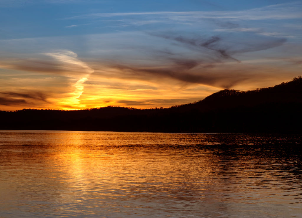 The sunset paints the sky over the hills of the Ohio River Valley with the reflection of fading evening light upon the calm water as photographed from the West Virginia shore